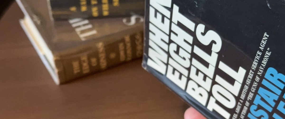 Hand holding When Eight Bells toll with a stack of books in the background